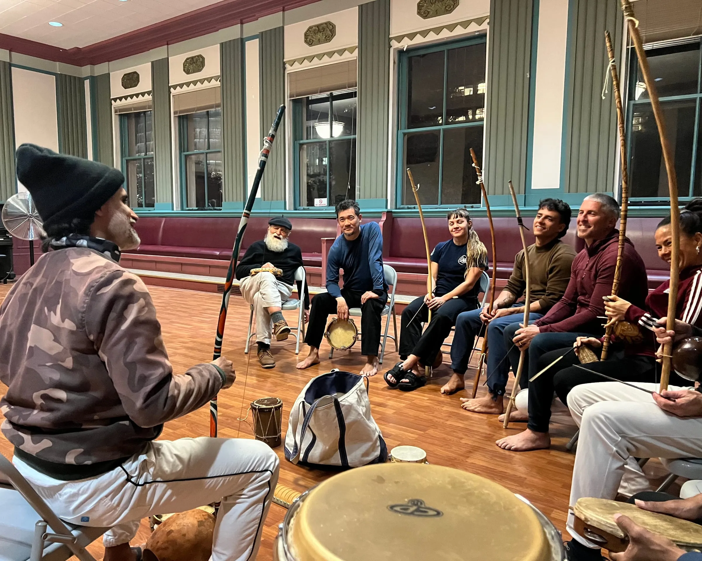 Students hold berimbaus in a music circle while Mestre Tico demonstrates in a spacious hall.