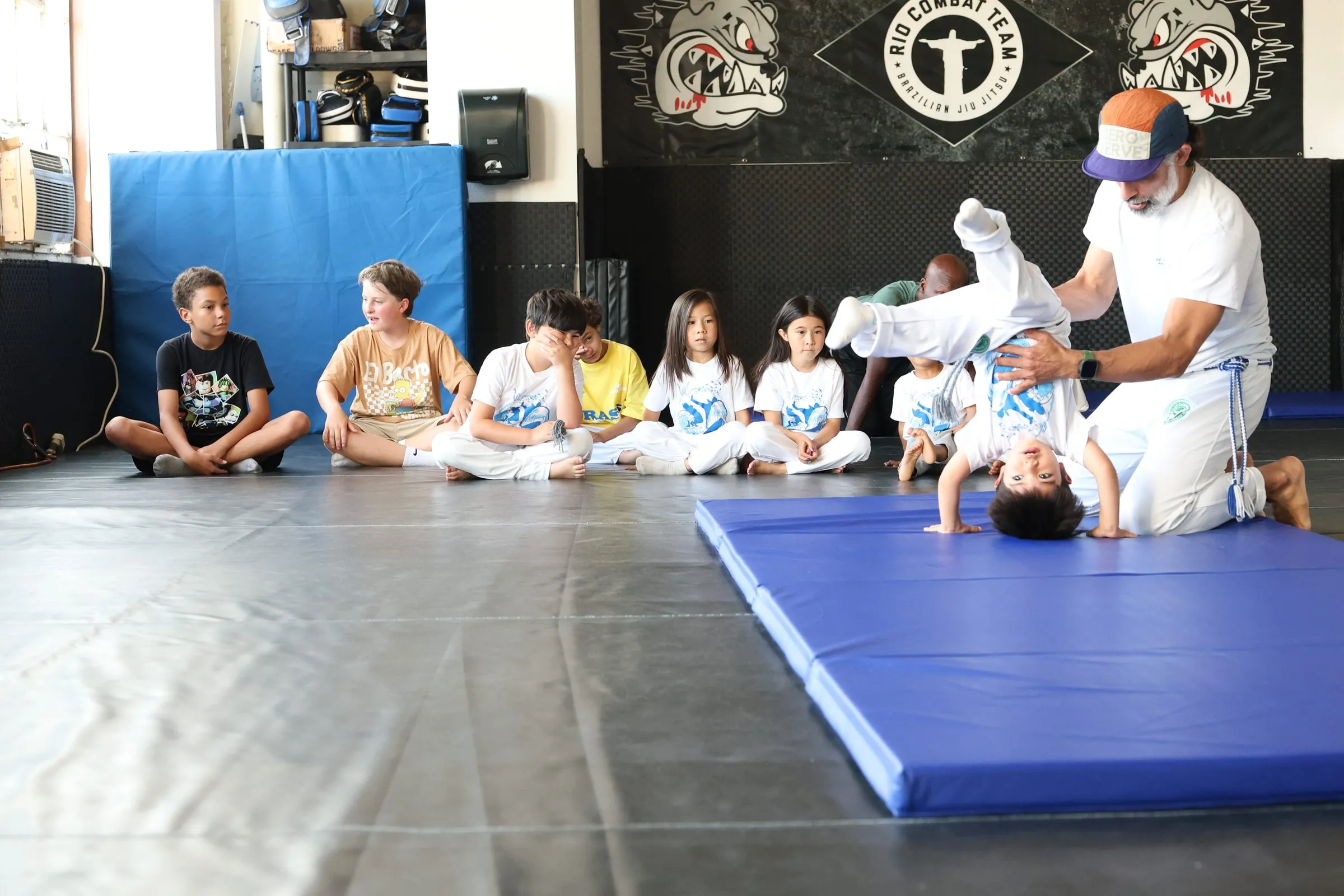 An instructor supports a child through a handstand while a group of children watch from the mat during a kids capoeira class.